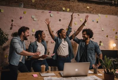 A startup team celebrating with raised arms around a laptop, sticky notes on the wall